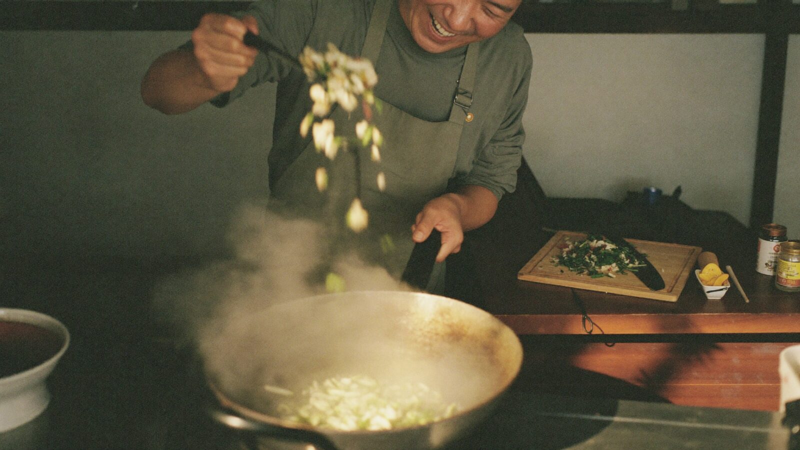 A man laughing as he tosses vegetables in a wok in a small home kitchen