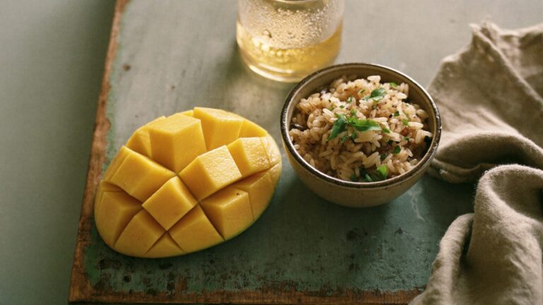 A halved fresh mango and a bowl of rice on a timber board beside a glass of iced tea