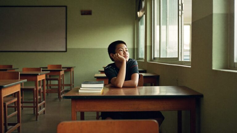 A teenager sitting alone at a desk in an empty classroom, looking out of the window