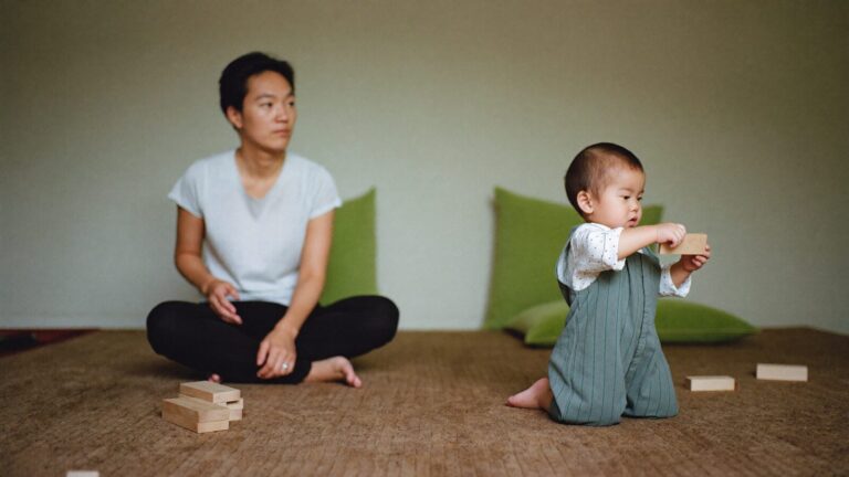 A parent watching with quiet concern as their young child plays alone with wooden blocks