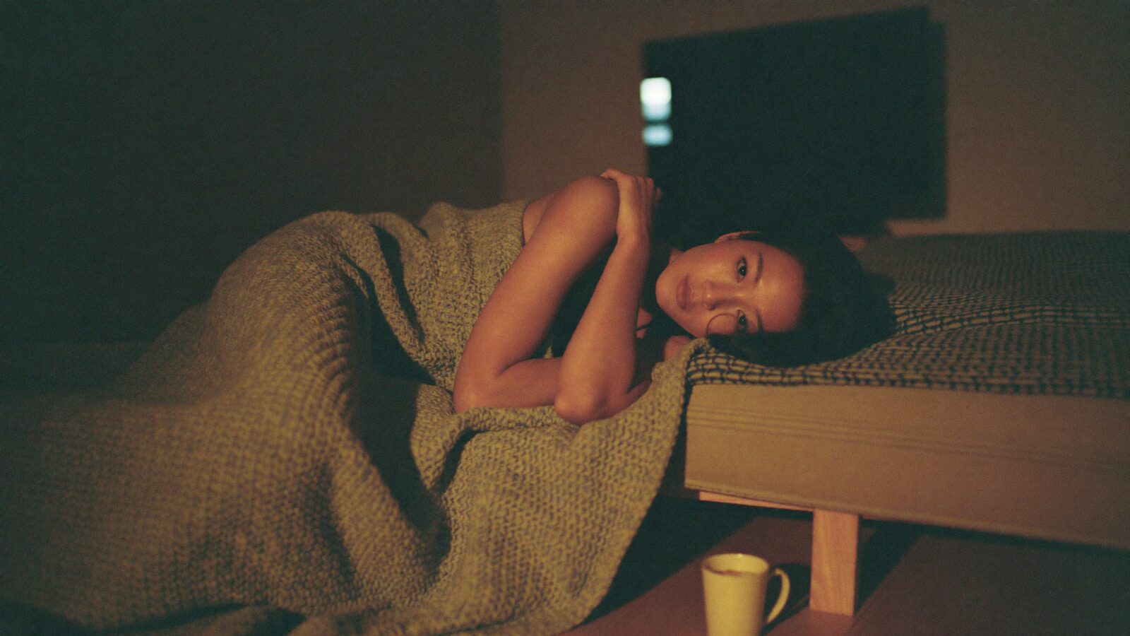 A woman curled under a blanket on a sofa watching television in the evening