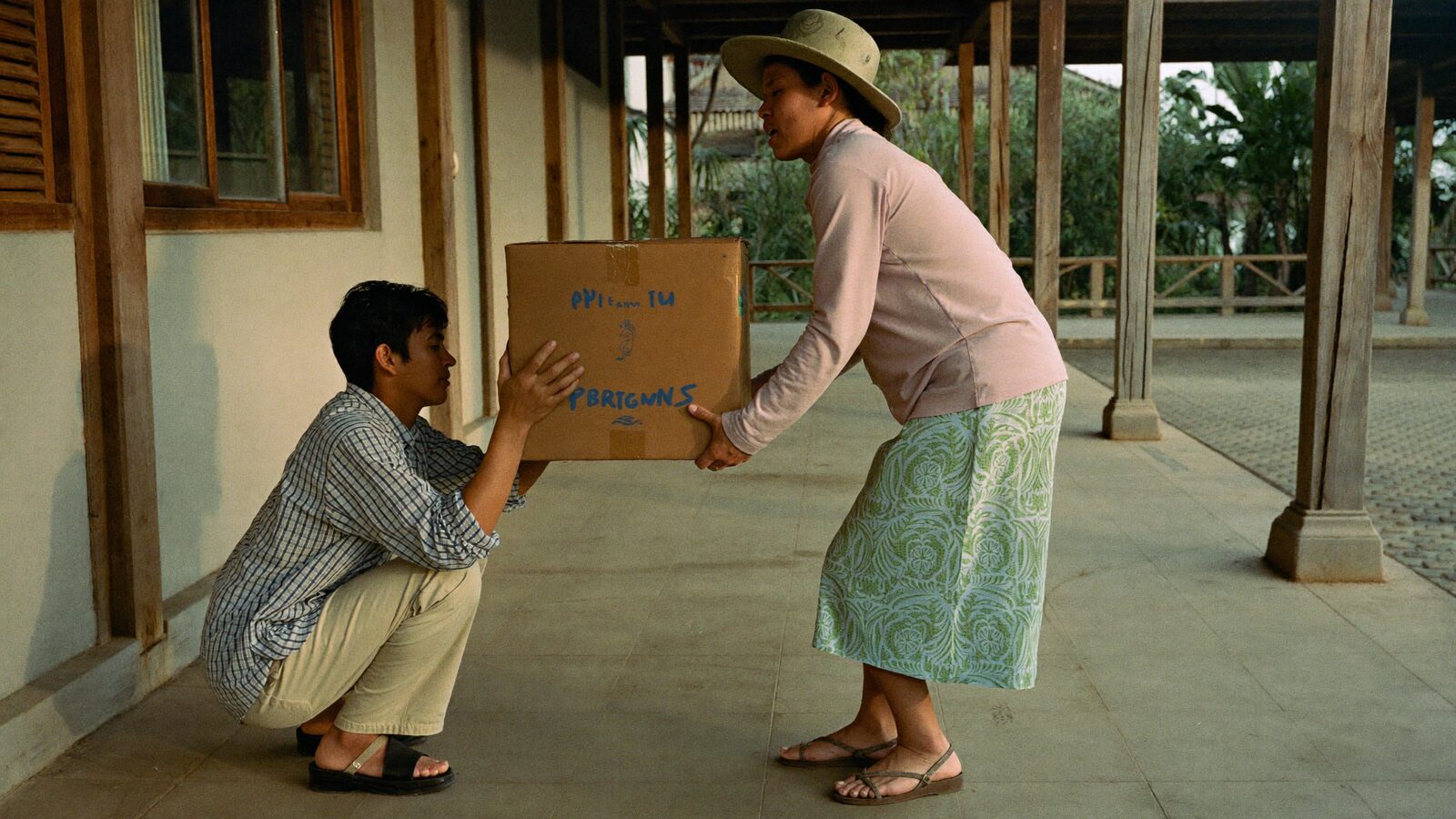 A young man steadying a heavy box for an older woman on a covered walkway