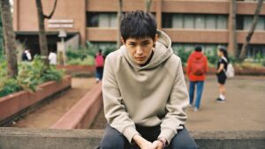 A young adult sitting alone on a low wall outside a college, shoulders rounded