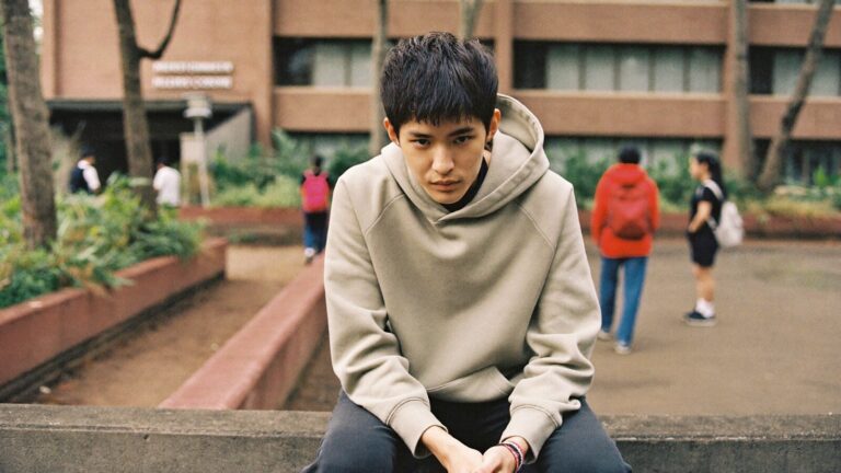 A young adult sitting alone on a low wall outside a college, shoulders rounded