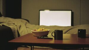 A coffee table with a remote control, snacks and mugs lit by the glow of a television
