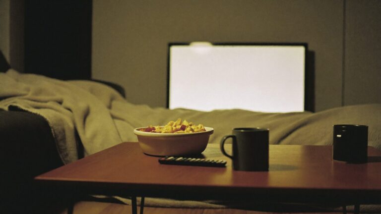 A coffee table with a remote control, snacks and mugs lit by the glow of a television